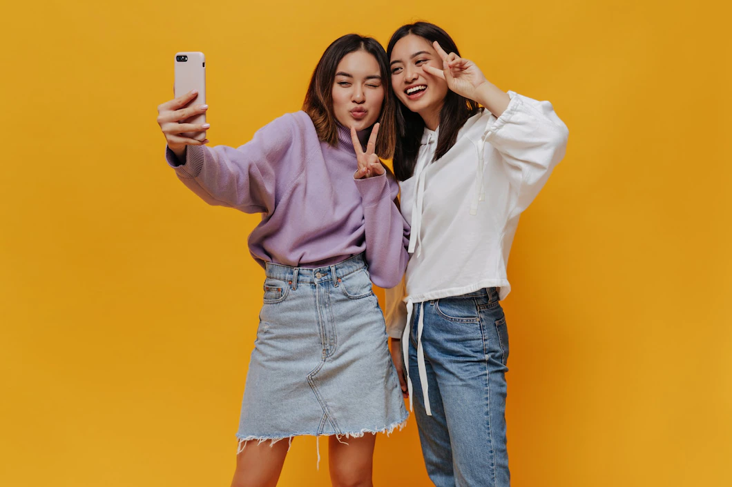  Two young women taking a selfie with a smartphone, isolated over a yellow background.